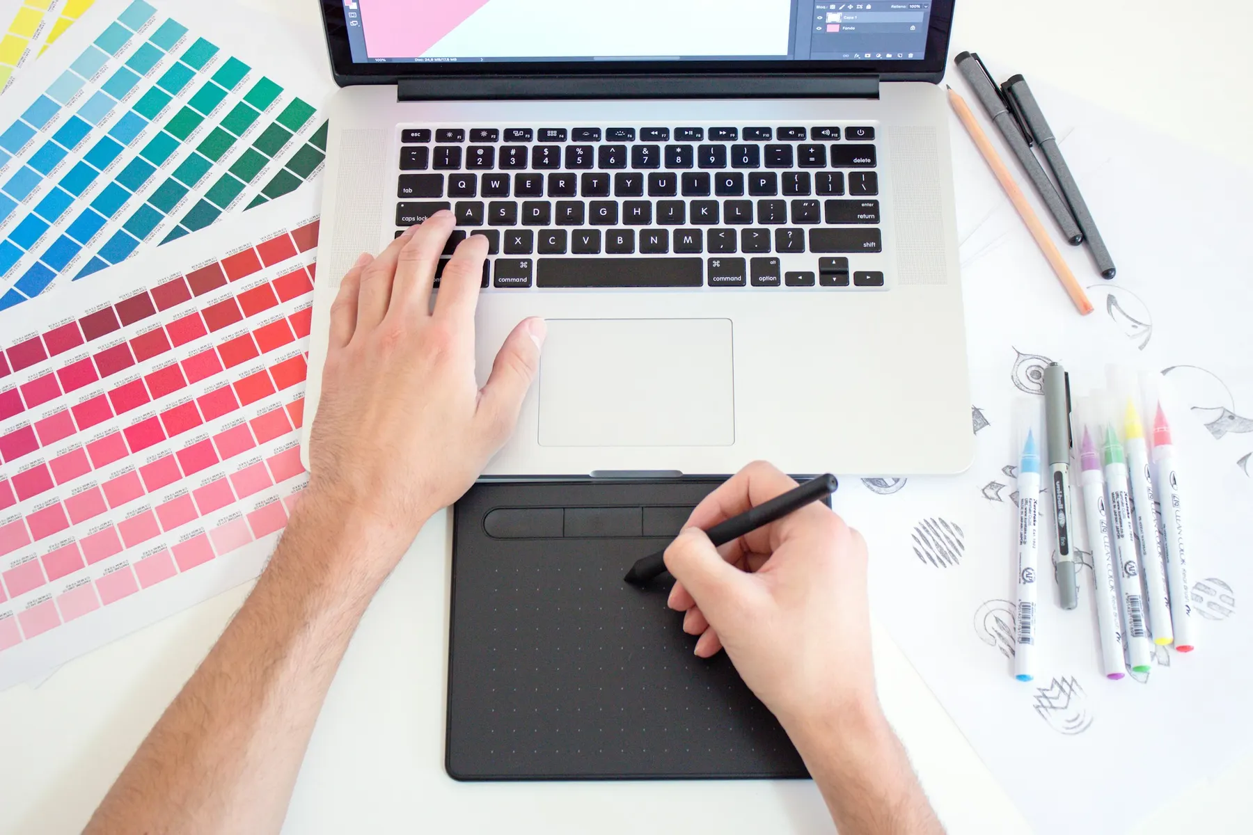 Une main dessine sur une tablette graphique à côté d’un ordinateur portable et de nuanciers